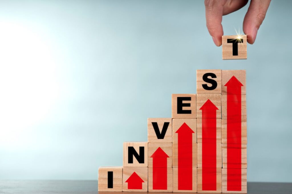 Mão posicionando blocos de madeira formando a palavra INVEST em escada crescente com setas vermelhas representando crescimento de investimentos