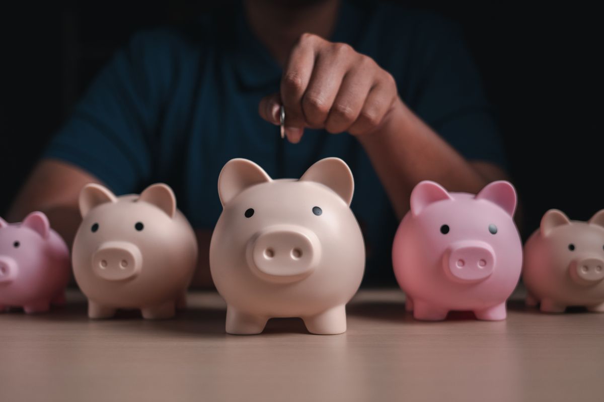 Como começar a investir: mão colocando moeda em cofrinhos representando diferentes tipos de investimentos e diversificação financeira