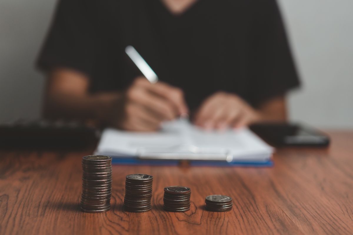 Pilhas decrescentes de moedas em primeiro plano sobre mesa de madeira, com pessoa ao fundo fazendo anotações em caderno e calculadora ao lado, ilustrando planejamento financeiro e controle de gastos